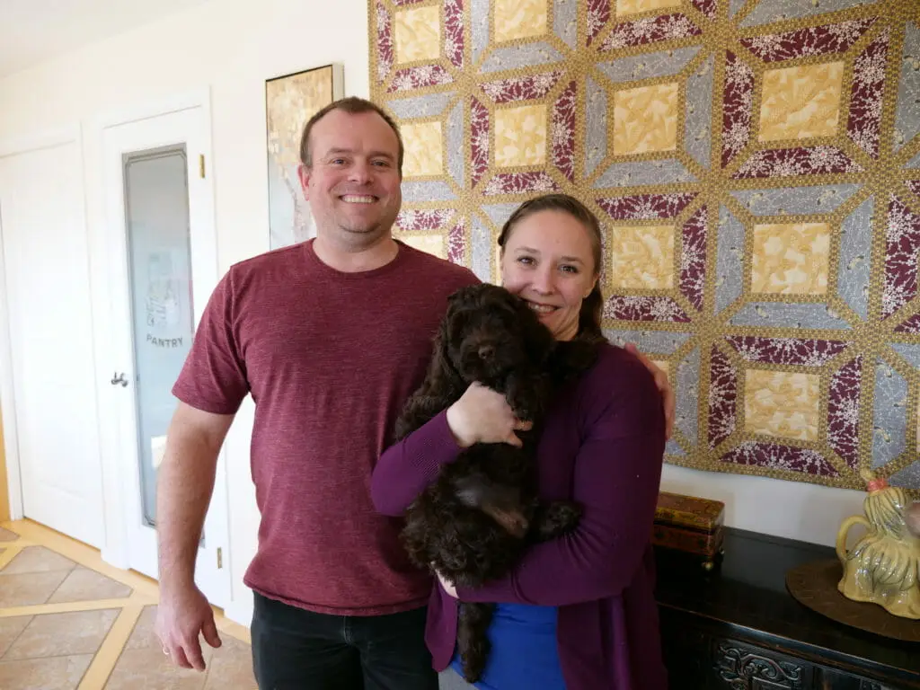 A grinning man stands next to a happy woman who is holding their new 8-week old brown labradoodle puppy. Behind them is a multicolored wall hanging.