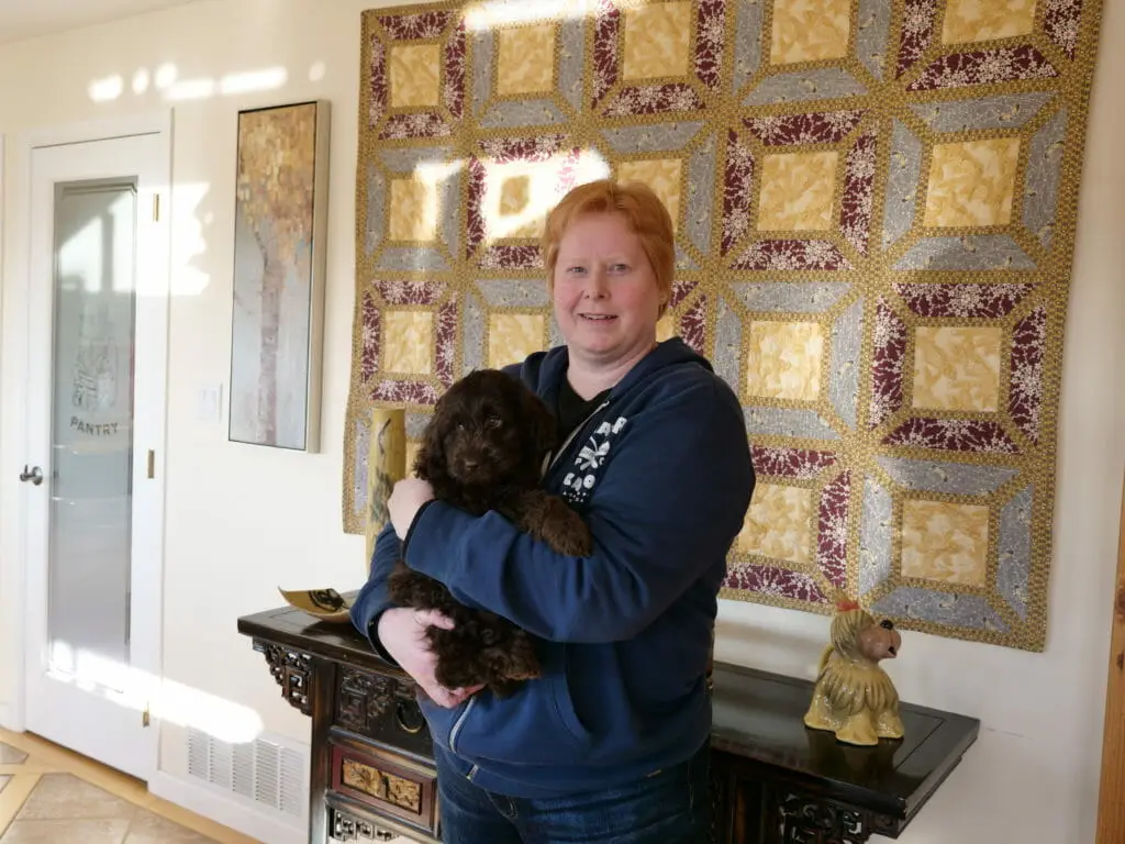 A smiling woman holds her new 8-week old brown labradoodle puppy in front of a multicolored wall hanging