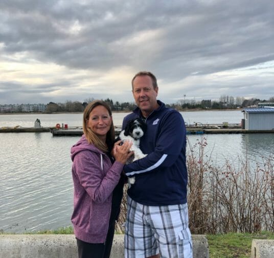 A couple standing in front of a body of water, holding their new 8-week old black and white labradoodle puppy. They are smiling.