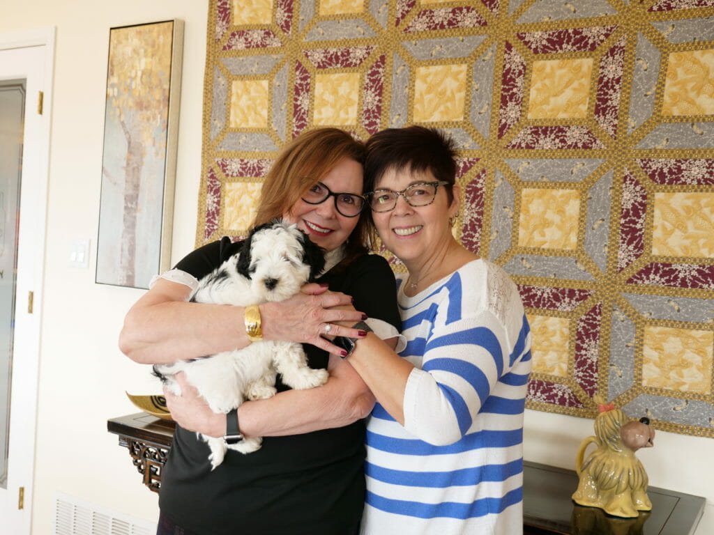 2 women smile brightly as they cuddle their new 8-week old black and white labradoodle puppy.