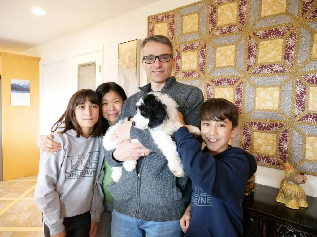 A man is grinning while holding a black and white labradoodle puppy (8-weeks old) surrounding him, with big smiles are his wife and 2 children.