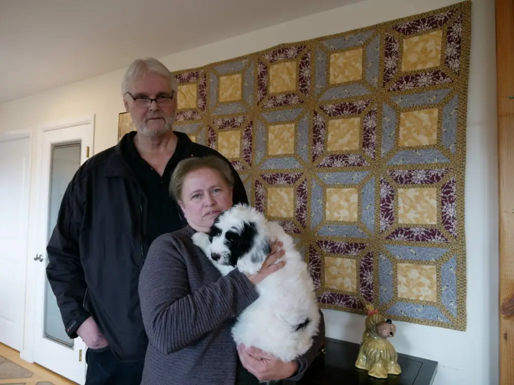 2 adults standing in front of a multicolored wall hanging, the woman is holding their new 8-week old black and white labradoodle puppy.