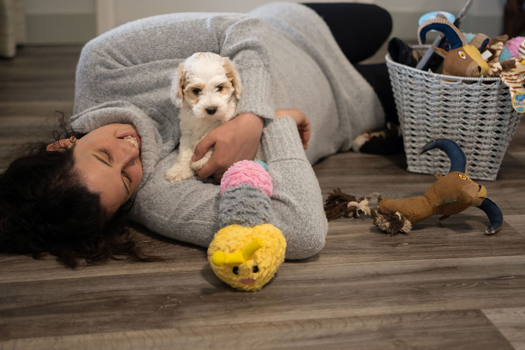 a woman is laying down on the floor with a puppy in her arms. She has her head towards the camera and the puppy is looking towards the camera. A basket of toys is beside them